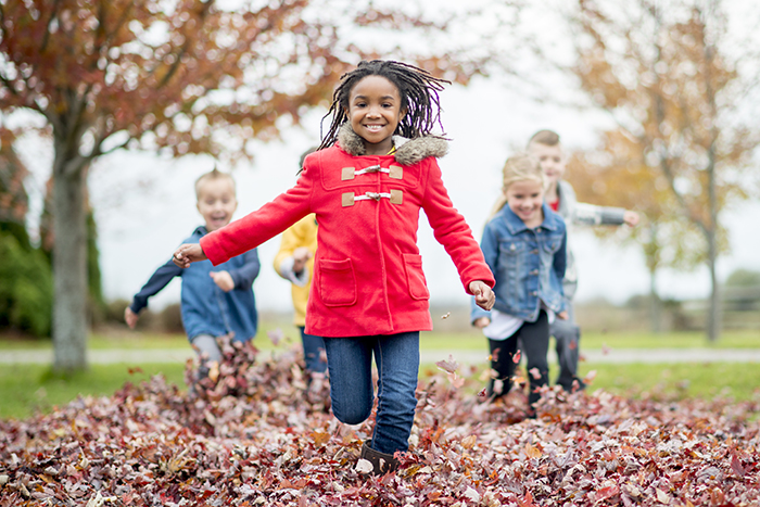 A multi-ethnic group of elementary age children are running happily through a pile of leaves on a beautiful fall day. One little girl is smiling and looking at the camera.